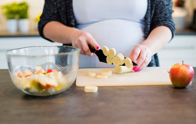 Gravid kvinna som skär frukt på en skärbräda på en köksbänk. Den skurna frukten är placerad i en glasskål vid sidan av skärbrädan.