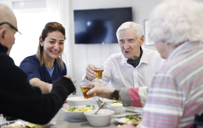 Tre äldre och en yngre person äter mat vid ett bord . De skålar med varandra och ser glada ut.