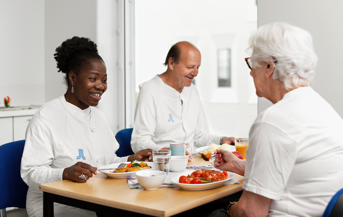Tre patienter sitter och äter tillsammans i ett dagrum på ett sjukhus. De ser glada ut och pratar med varandra.