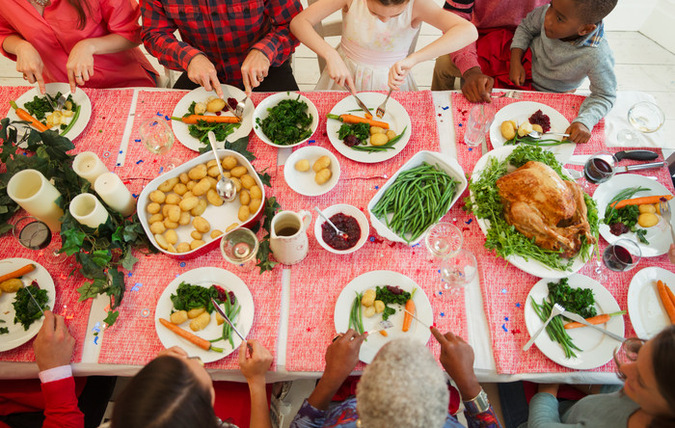 En bild ovanifrån där en grupp människor i olika åldrar äter en middag vid ett dukat bord. På bordet finns det mat på både fat och tallrikar. Maten består av potatis, böner, morötter, sås, gelé och en stor kalkon.