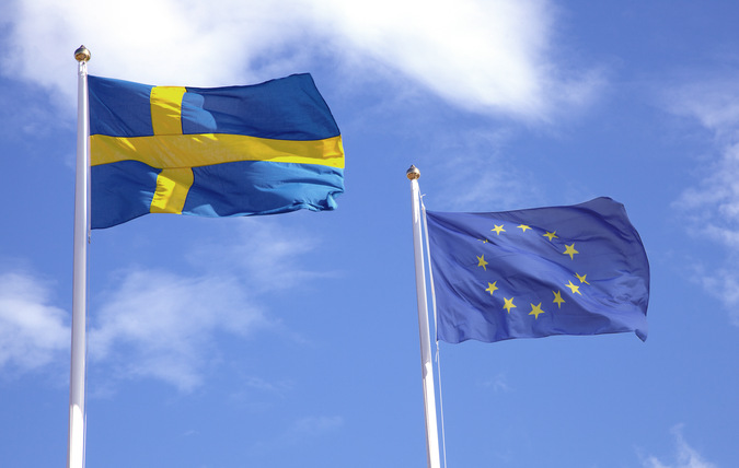 Två flaggstänger med en svenska flagga och en EU flagga, blå himmel med vita moln i bakgrunden 