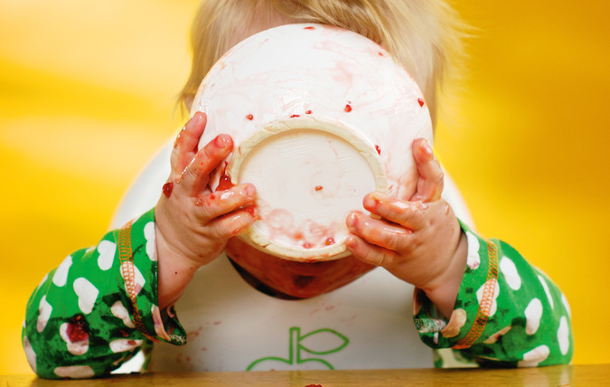 Spädbarn äter mat ur en skål, skålen täcker hela ansiktet och barnets händer är kladdiga.