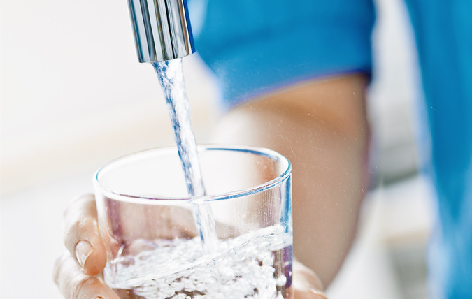 Ett dricksglas som fylls med vatten från en vattenkran. Glaset hålls av en person med blå tröja, bara armen och handen syns i bild.