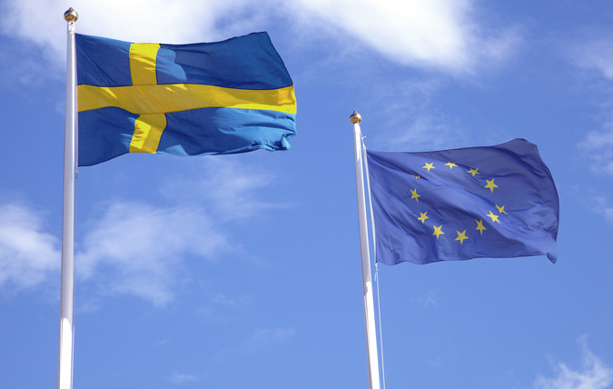 Två flaggstänger med en svenska flagga och en EU flagga, blå himmel med vita moln i bakgrunden 