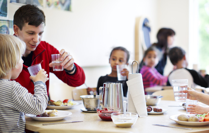 Fyra barn äter tillsammans med en förskolepedagog vid ett bord i en matsal.