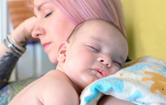 Ett barn som sover på en kvinnas axel, kvinnan blundar också. På axeln har kvinnan en filt med figurer som barnet har som kudde.