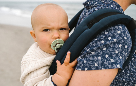 Närbild på en bebis i en bärsele på ryggen vid havet.