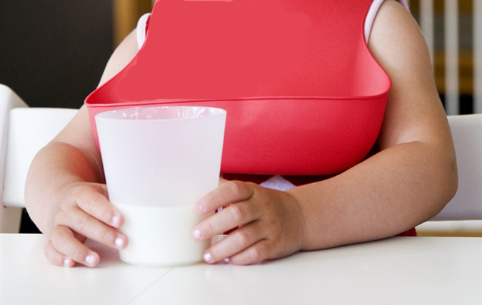 Barnhänder håller i ett glas mjölk. I bakgrunden syns en haklapp. 