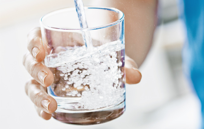Ett dricksglas som fylls med vatten från en vattenkran. Glaset hålls av en person med blå tröja, bara armen och handen syns i bild.